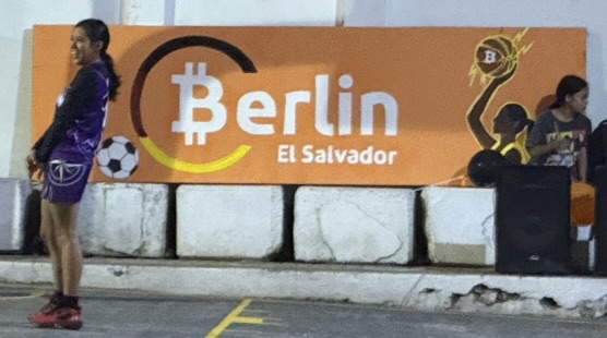 The completed orange Berlín El Salvador sign displayed at the basketball court, with a football on the left and a basketball player on the right.