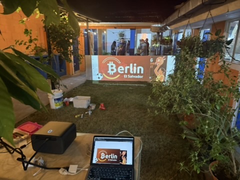 The Berlín sign being painted in the community center courtyard, with a projector, laptop and paint supplies in the foreground.