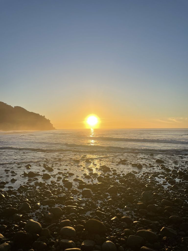 Photograph of sunrise over the Pacific at Punta El Zonte, El Salvador: the sun sits on the horizon beside a dark headland on the left, with smooth wet stones covering the beach in the foreground.